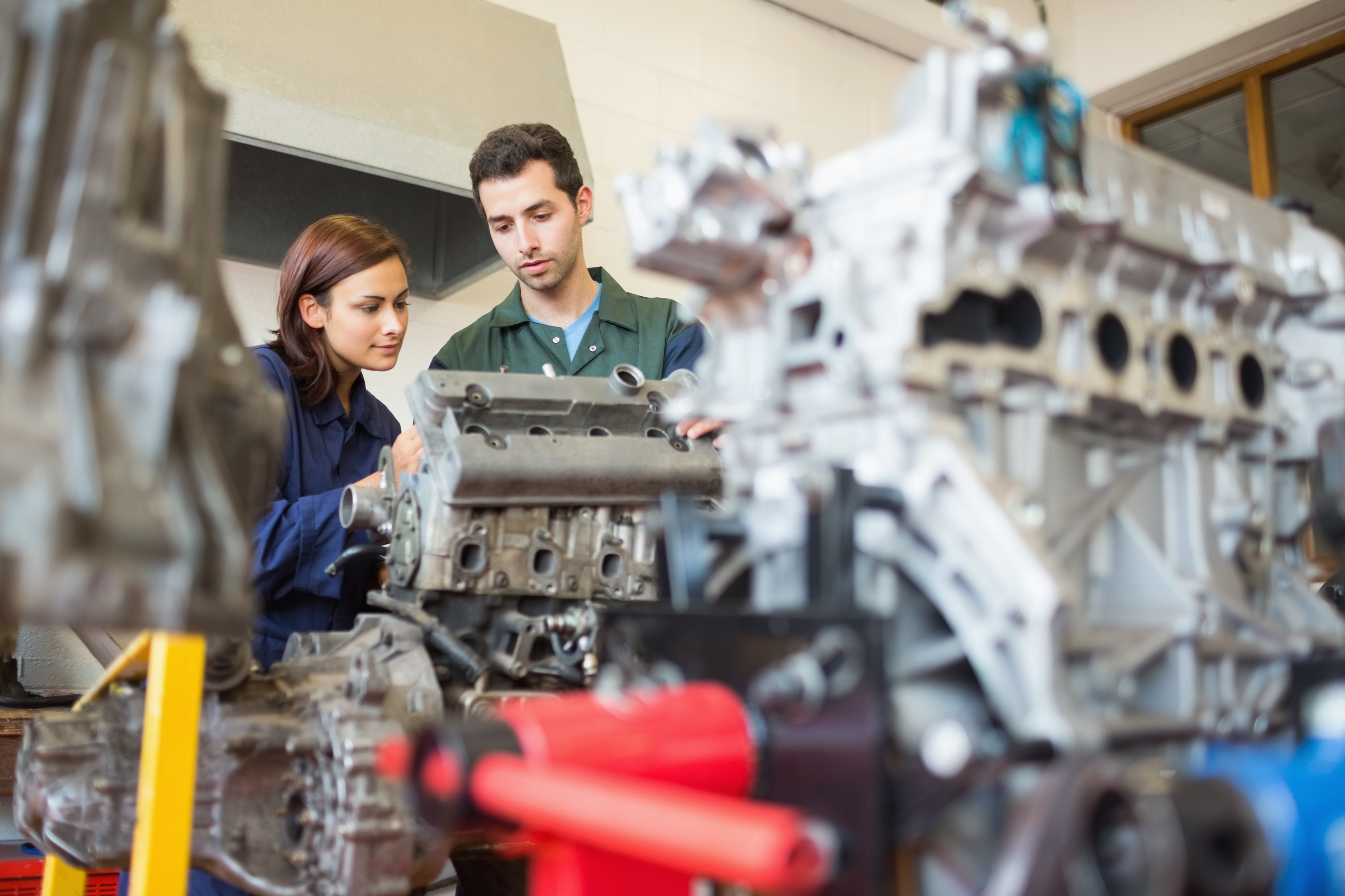 Zwei Personen inspizieren Automotor, im Vordergrund verschwommen weitere Motorenteile