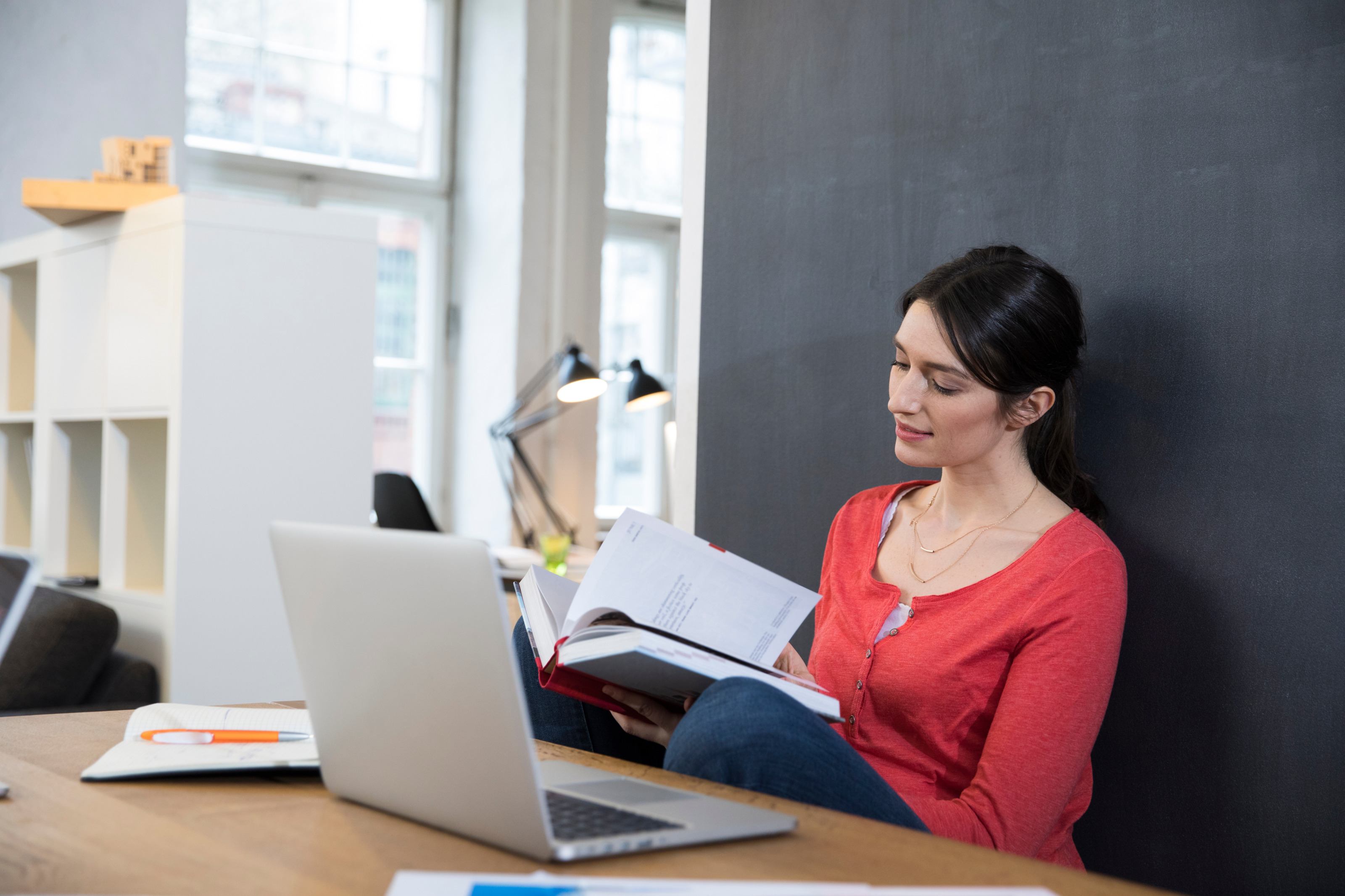 Eine Person sitzt hinter einem Tisch und lehnt sich an einer dunklen Wand an. In den Händen hält sie ein aufgeklapptes Buch, auf das sie blickt. Vor ihr auf dem Tisch steht ein aufgeklappter Laptop